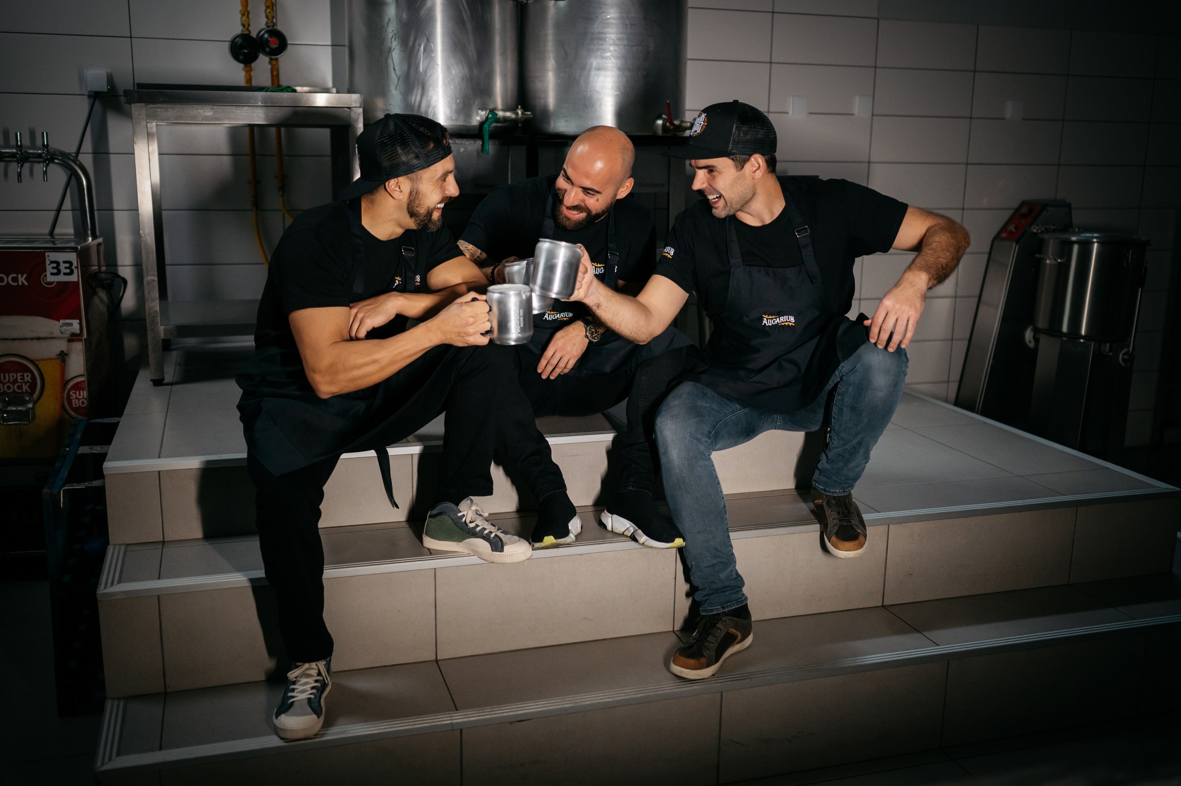 Os três sócios do Alicarius sentados na cozinha, a brindar com canecas de cerveja