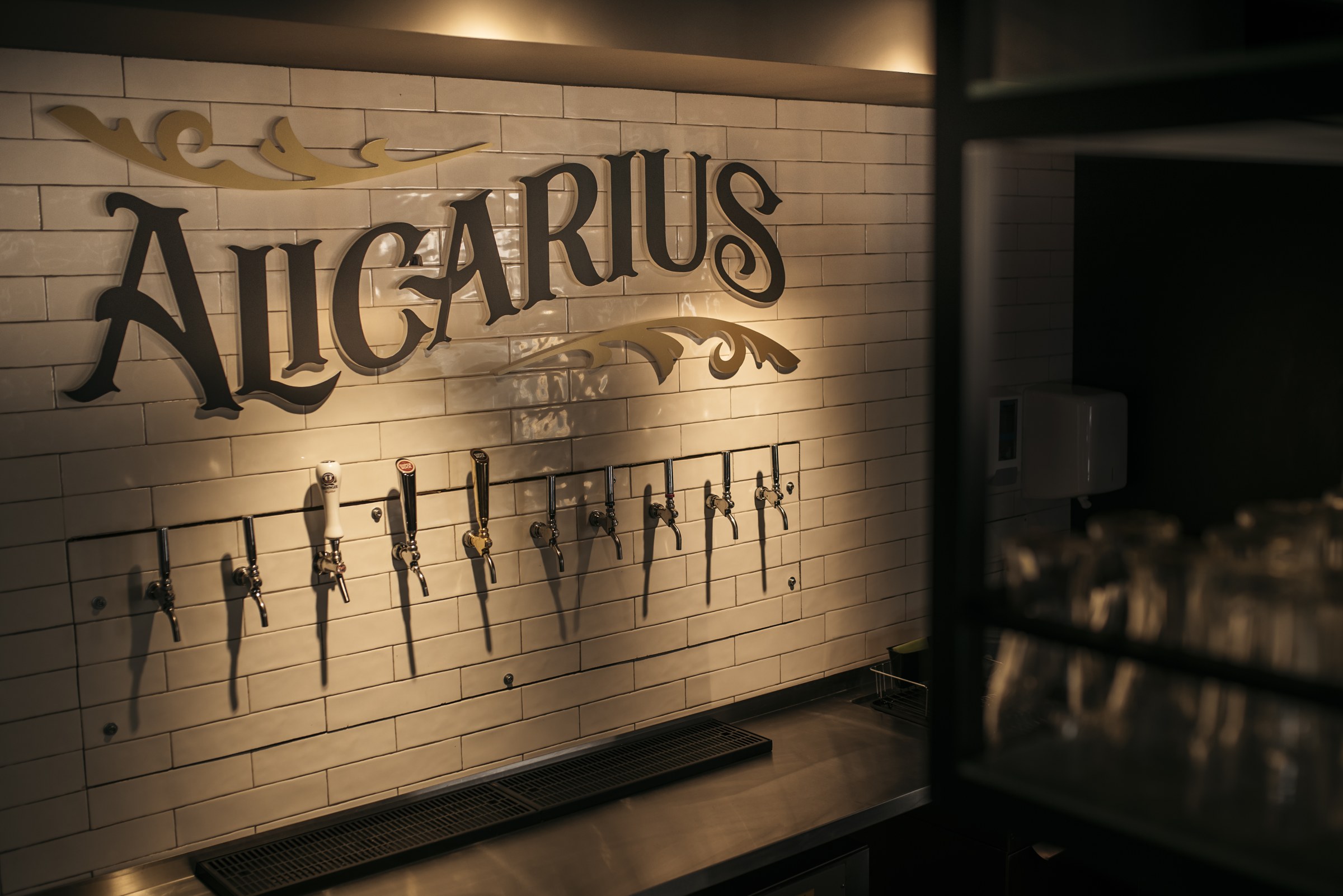 Parede de azulejo branco com o letreiro Alicarius e as torneiras de cerveja