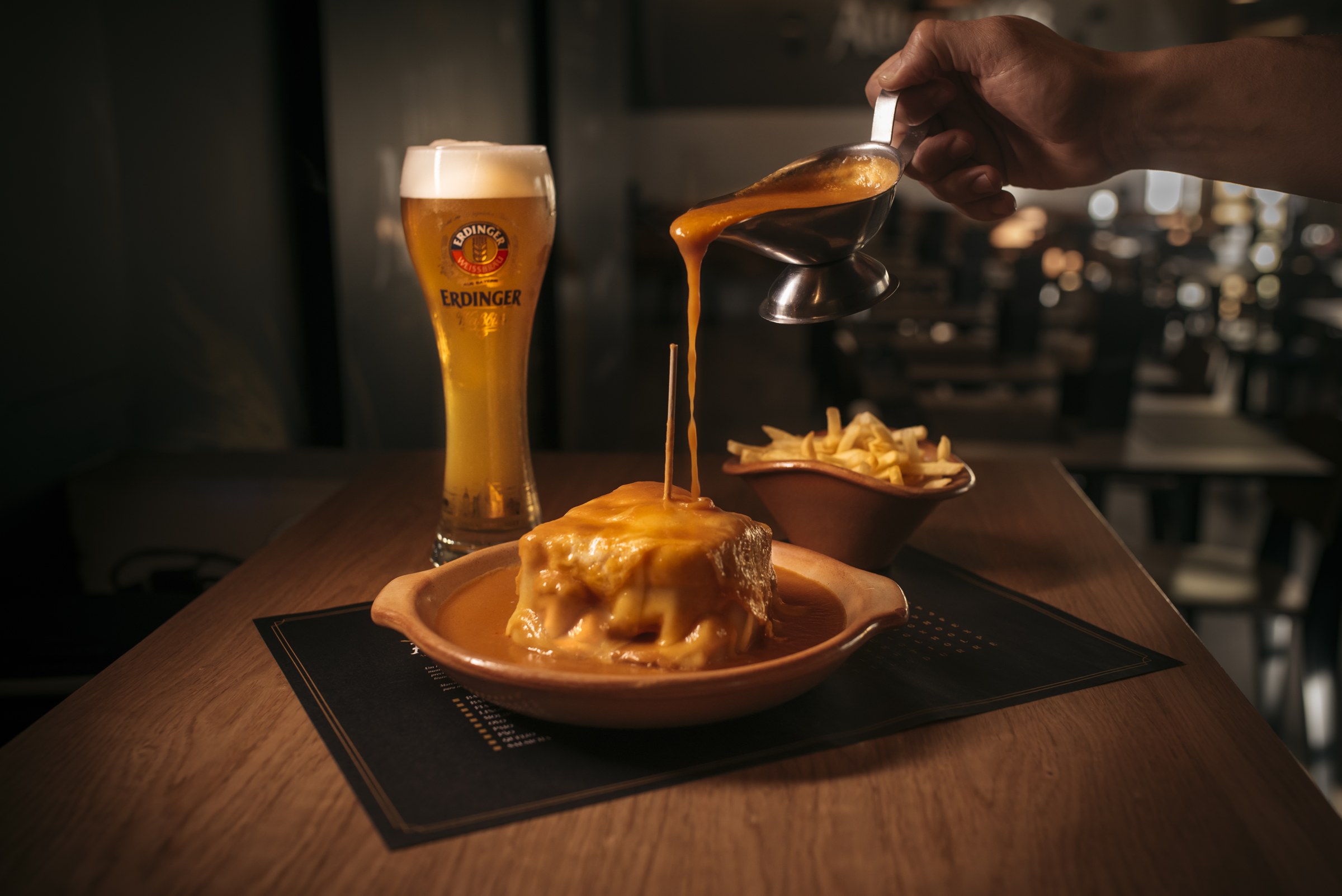 Molho a ser deitado sobre uma francesinha, com cerveja e batatas fritas ao lado
